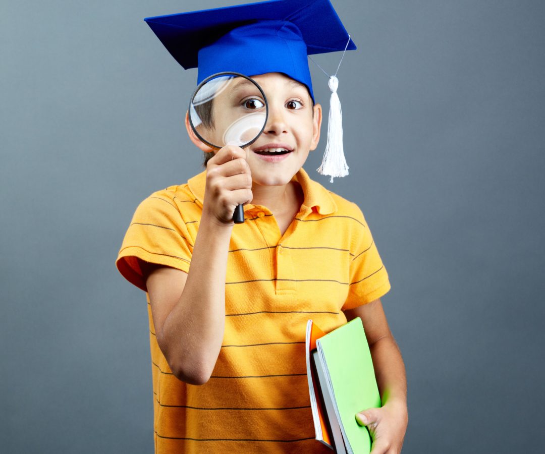 Portrait of curious boy in graduation hat looking through magnifying glass